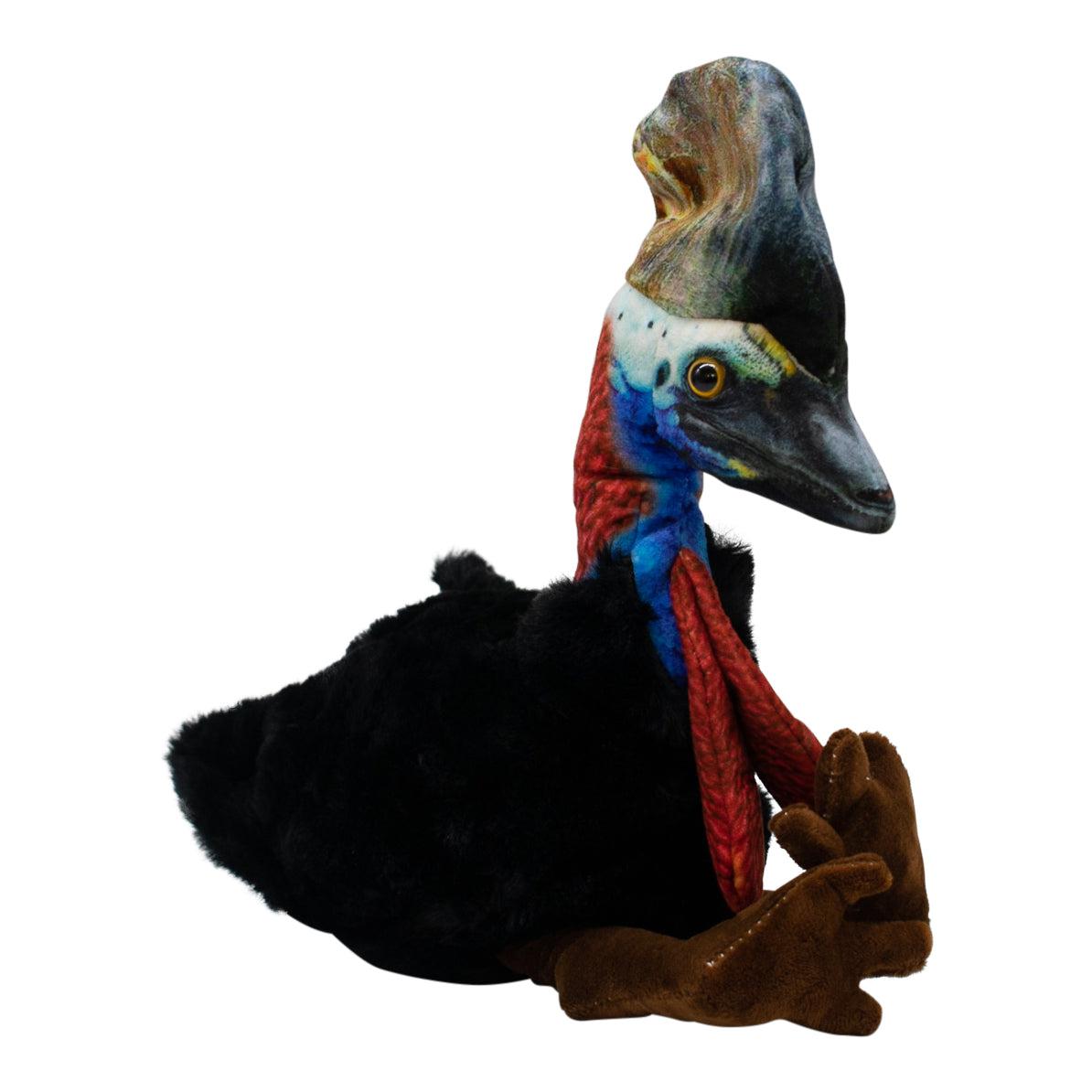 Lifelike Kay Cassowary plush toy sitting, with black furry body, red and blue neck, yellow eyes, and a colorful textured casque.